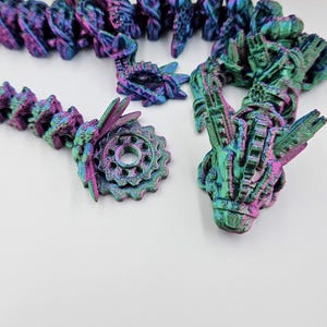 May include: A close-up of a multi-colored iridescent 3D printed dragon figurine with a gear-shaped base. The dragon has a detailed, intricate design and is made of a shiny, metallic material.