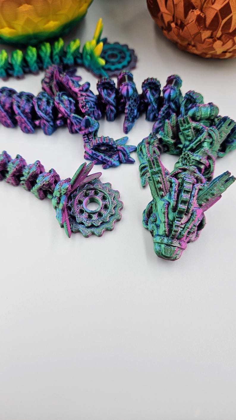 May include: A close-up of a multi-colored iridescent 3D printed dragon figurine with a gear-shaped base. The dragon has a detailed, intricate design and is made of a shiny, metallic material.