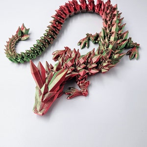 May include: A 3D printed dragon figurine with a red and green crystal-like design. The dragon is curled up in a resting position with its head resting on the ground.