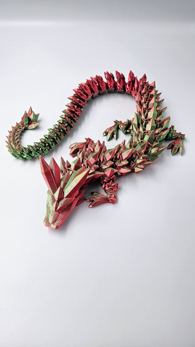 May include: A 3D printed dragon figurine with a red and green crystal-like design. The dragon is curled up in a resting position with its head resting on the ground.