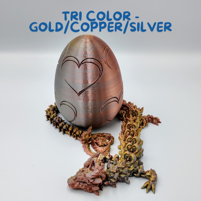 May include: A tri-color dragon egg with a gold, copper, and silver finish. The egg is decorated with hearts and circles. The dragon is curled up around the egg.