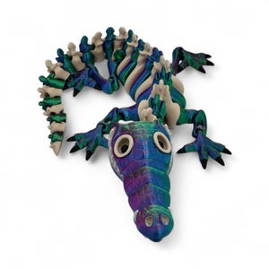 Pu&ograve; includere: Scultura stilizzata di un alligatore dai colori vivaci. Il corpo &egrave; blu, verde e viola, con una spina dorsale color crema. Gli occhi sono grandi e bianchi, e gli artigli neri. L'opera &egrave; posta su uno sfondo bianco, che ne esalta i colori.