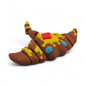 May include: A brown croissant-shaped object with yellow and red accents, resembling butter and jam. The pastry has a textured surface and two blue circular details. The object is set against a white background.