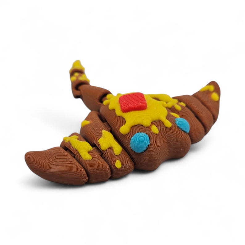 May include: A brown croissant-shaped object with yellow and red accents, resembling butter and jam. The pastry has a textured surface and two blue circular details. The object is set against a white background.