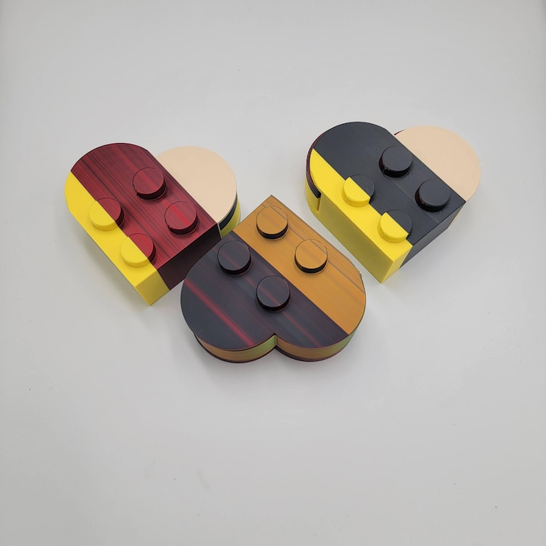 May include: Three heart-shaped boxes in a playful design, resembling building blocks. Each box features a unique color scheme: red and yellow, black and yellow, and brown and orange. The boxes have a smooth, polished finish.