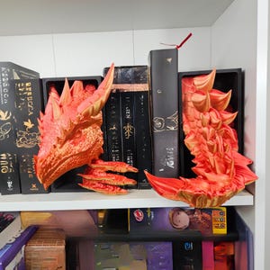 May include: Red and gold dragon-themed bookends on a white shelf. The bookends are 3D-printed and feature a dragon head and tail. Books with black covers are placed between the bookends. The shelf is part of a white bookcase.