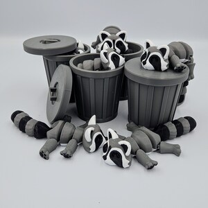 May include: Gray 3D-printed raccoon toys with black and white markings are scattered around several gray trash cans. Some raccoons are inside the cans, while others are lying nearby. The trash cans have lids.