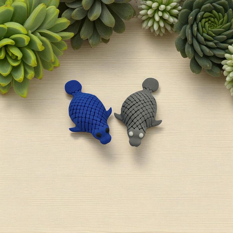 Puede incluir: Dos figuras con forma de foca. Una es azul, la otra gris. Ambas tienen un patr&oacute;n texturizado, similar a escamas, en la espalda y peque&ntilde;as aletas. Las figuras est&aacute;n sobre un fondo claro con vetas de madera, con suculentas verdes en las esquinas.