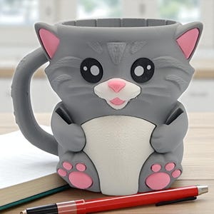 May include: A gray cat-shaped mug with pink accents on the ears, nose, and paws. The mug has a white belly and large black eyes. A red pen and a notebook are in the foreground.