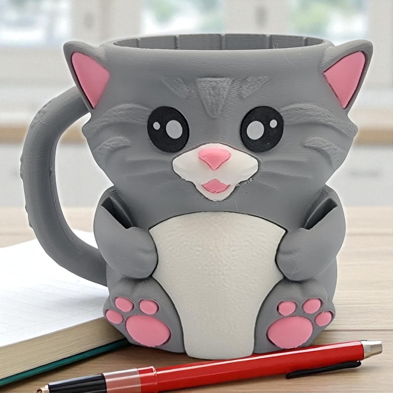 May include: A gray cat-shaped mug with pink accents on the ears, nose, and paws. The mug has a white belly and large black eyes. A red pen and a notebook are in the foreground.