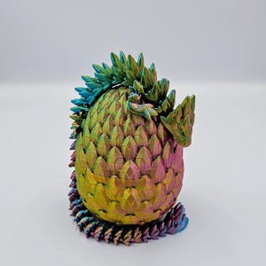 May include: A iridescent dragon egg figurine with a green, blue, and purple color scheme. The egg is made of a textured material that resembles scales. The dragon is curled up inside the egg.