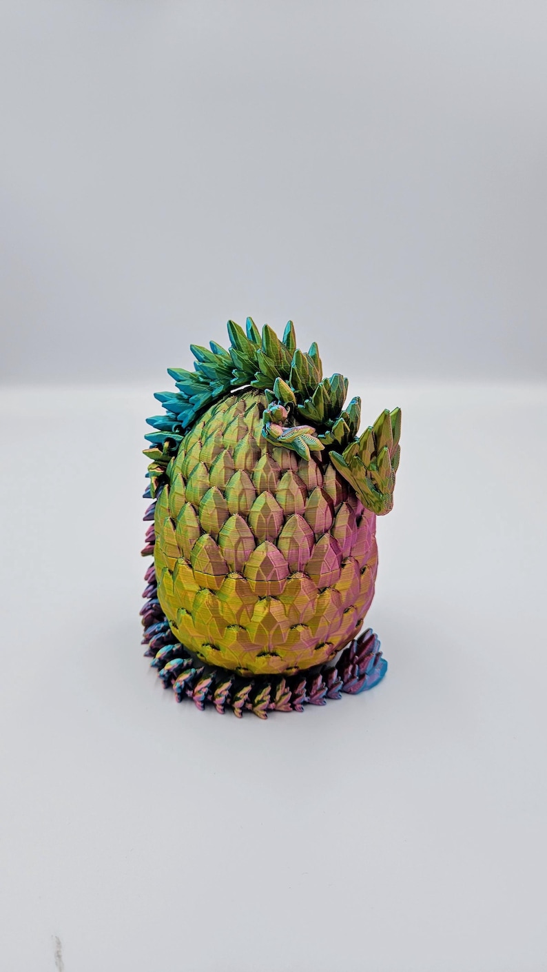 May include: A iridescent dragon egg figurine with a green, blue, and purple color scheme. The egg is made of a textured material that resembles scales. The dragon is curled up inside the egg.