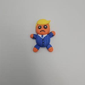 May include: A handmade voodoo doll with a yellow hairstyle, orange skin, and a blue suit. The doll's eyes are stitched with black Xs, and its mouth is stitched with a frown. The doll's hands and feet are orange.