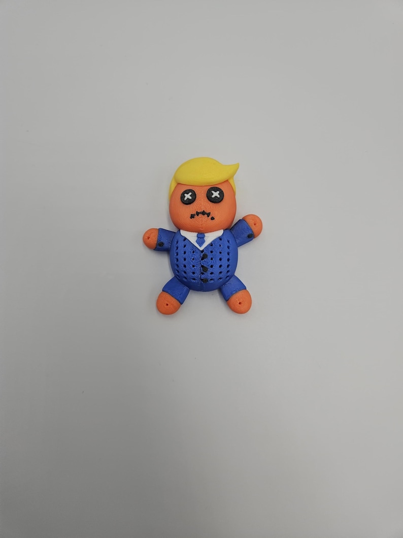 May include: A handmade voodoo doll with a yellow hairstyle, orange skin, and a blue suit. The doll's eyes are stitched with black Xs, and its mouth is stitched with a frown. The doll's hands and feet are orange.