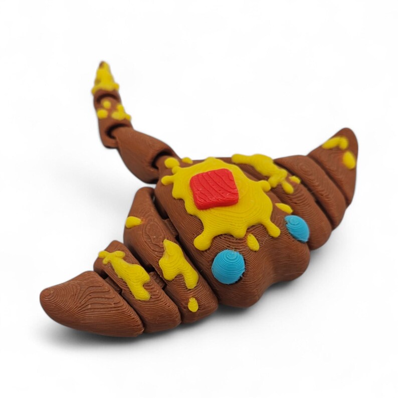 May include: A brown, croissant-shaped sculpture with yellow and red accents, and blue details. The pastry-like design features a textured surface and is decorated with what appears to be butter and a red square.