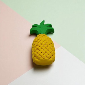 May include: A yellow pineapple with a green leafy top. The pineapple has a textured surface and is set against a backdrop of pink, green, and white geometric shapes.