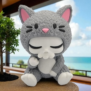 May include: A gray figurine of a cat-shaped character with a skull-like face. The cat has pink inner ears and a pink nose. The figurine is sitting on a woven surface, with a small tree and ocean in the background.