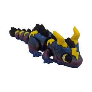K&ouml;nnte beinhalten: Eine 3D-gedruckte Drachenfigur mit einem Galaxie-Thema. Der Drache hat einen blauen K&ouml;rper mit lila und wei&szlig;en Sprenkeln, schwarzen Stacheln und gelben H&ouml;rnern und Akzenten. Die Augen sind gelb mit einer schwarzen Pupille.