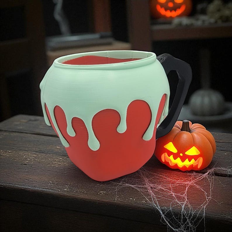 Pode incluir: Uma caneca vermelha e verde-menta em forma de caldeir&atilde;o com uma al&ccedil;a preta, cheia de l&iacute;quido vermelho. Uma pequena lanterna de ab&oacute;bora iluminada fica ao lado da caneca. A caneca tem um design gotejante, perfeito para o Halloween.