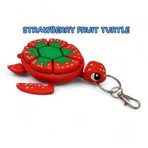 May include: A red and green strawberry-shaped turtle keychain. The turtle's shell is made up of individual strawberry slices with white seeds. The turtle has a green head and a silver key ring.