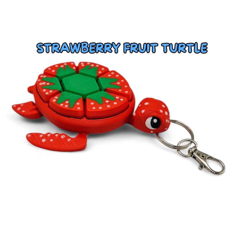 May include: A red and green strawberry-shaped turtle keychain. The turtle's shell is made up of individual strawberry slices with white seeds. The turtle has a green head and a silver key ring.