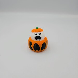 May include: A small, white ghost figurine wearing an orange pumpkin costume with the word "BOO" printed on the front.