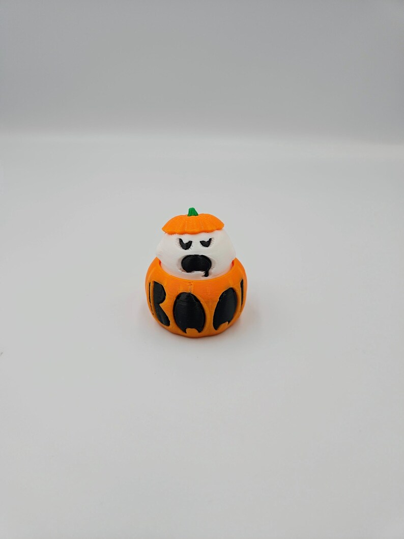 May include: A small, white ghost figurine wearing an orange pumpkin costume with the word "BOO" printed on the front.
