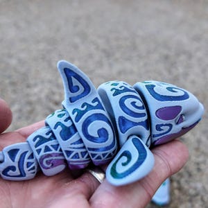 May include: A light blue, segmented shark toy with a blue and green tribal pattern. The toy is made of plastic and has a segmented body.