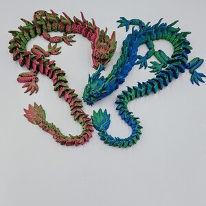 May include: Three articulated dragon figurines in shades of green, pink, and blue. The dragons have detailed scales and are posed in a curved position. The background is plain white.
