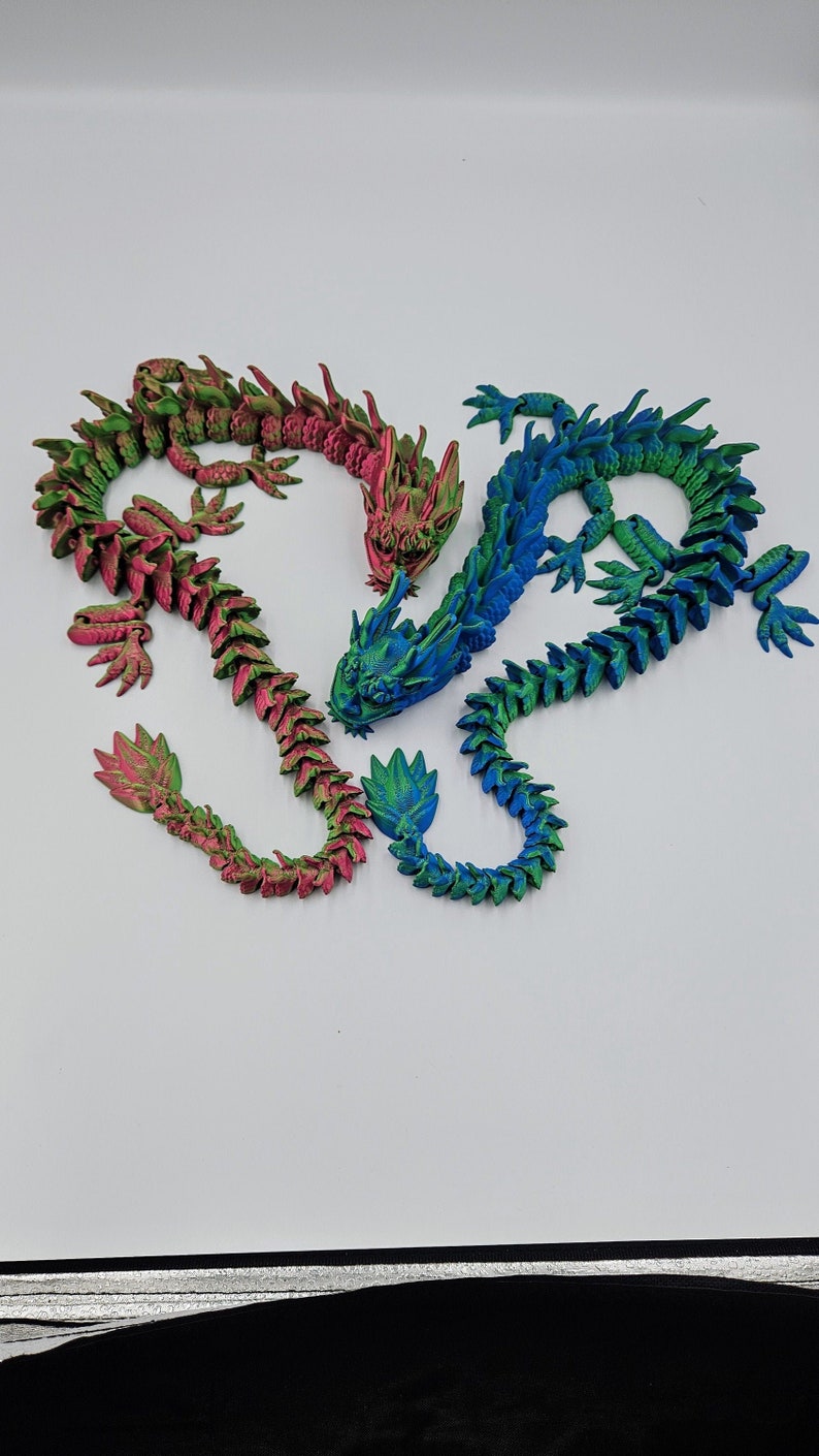 May include: Three articulated dragon figurines in shades of green, pink, and blue. The dragons have detailed scales and are posed in a curved position. The background is plain white.