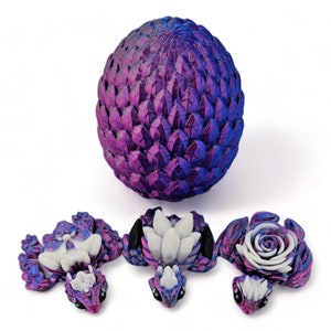 Puede incluir: Un huevo de drag&oacute;n morado y azul con tres dragones beb&eacute;s. Los dragones beb&eacute;s son blancos y tienen ojos negros. El huevo est&aacute; hecho de un material texturizado que parece escamas.