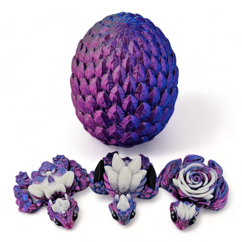 Puede incluir: Un huevo de drag&oacute;n morado y azul con tres dragones beb&eacute;s. Los dragones beb&eacute;s son blancos y tienen ojos negros. El huevo est&aacute; hecho de un material texturizado que parece escamas.