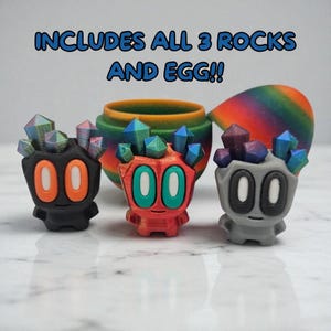 May include: Three colourful, stylised figures with crystal-like tops and large oval eyes. The figures are black, red, and grey, with orange, green, and white eye details. A rainbow-coloured egg and a small pot are in the background. Text reads: INCLUDES ALL 3 ROCKS AND EGG!!