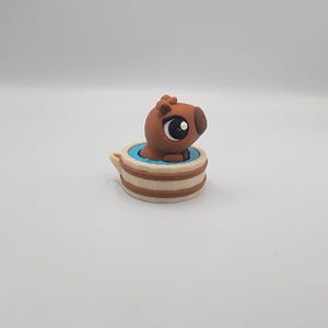 May include: A brown capybara figurine in a small, beige and brown tub with a blue interior. The capybara has large, dark eyes and is partially submerged in the blue water. The tub has a small handle.