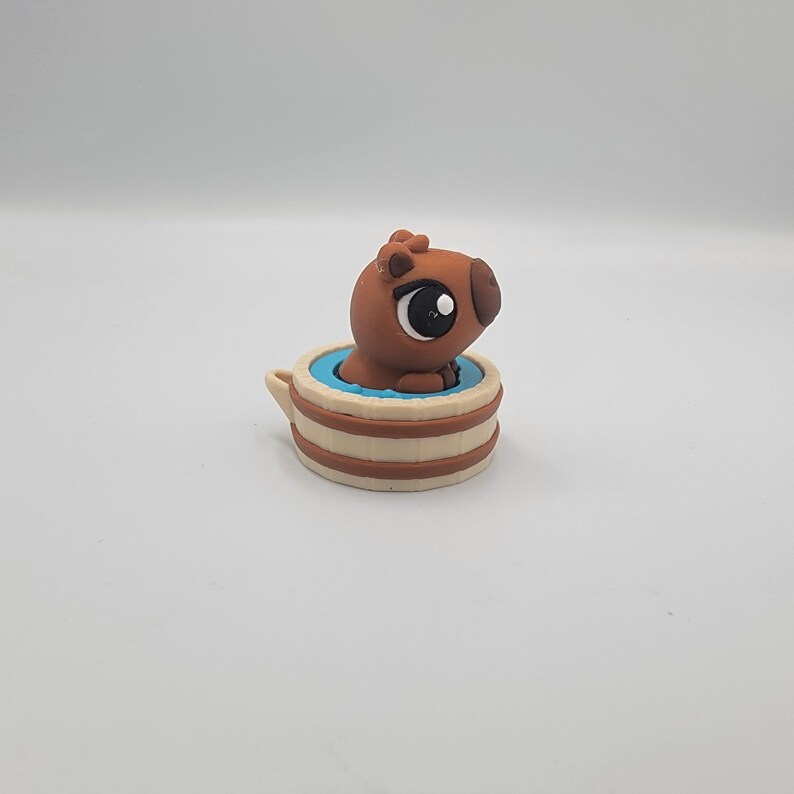May include: A brown capybara figurine in a small, beige and brown tub with a blue interior. The capybara has large, dark eyes and is partially submerged in the blue water. The tub has a small handle.