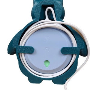 May include: A teal blue 3D printed wall mount for a smart speaker. The mount is shaped like a cartoon penguin and has a hole in the back for the speaker's power cord. The mount is designed to hold the speaker and its cord neatly.