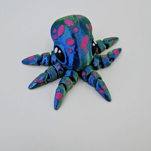 Puede incluir: Un peque&ntilde;o pulpo azul y verde con manchas rosas y ojos negros. El pulpo tiene ocho tent&aacute;culos y est&aacute; hecho de un material impreso en 3D.
