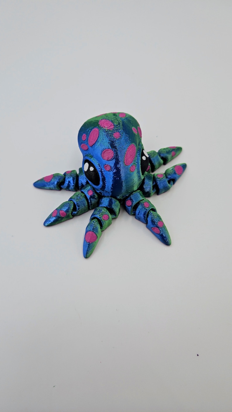 Puede incluir: Un peque&ntilde;o pulpo azul y verde con manchas rosas y ojos negros. El pulpo tiene ocho tent&aacute;culos y est&aacute; hecho de un material impreso en 3D.