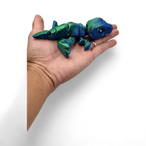 May include: A small, blue and green articulated dinosaur toy. The dinosaur is posed in a walking position with its head turned to the side. The toy is made of plastic and has a smooth, shiny finish.