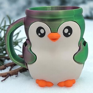 May include: A 3D-printed mug shaped like a penguin, with a white body, black eyes, and an orange beak and feet. The mug has a green and purple rim and handle, and is sitting in the snow.