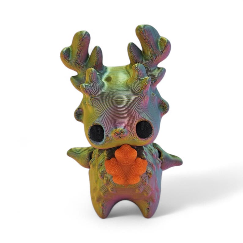 May include: A colorful, 3D-printed figurine of a deer-like creature with large black eyes and an orange chest emblem. The figure has a textured, layered appearance with a gradient of colors including green, yellow, purple, and blue. The figurine has antlers and small arms.