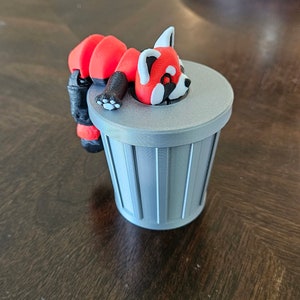 May include: A red panda figurine with white accents rests on a small, gray trash can. The panda has a black nose and paws. The trash can has vertical lines and a flat lid. The figurine is a whimsical decorative item.