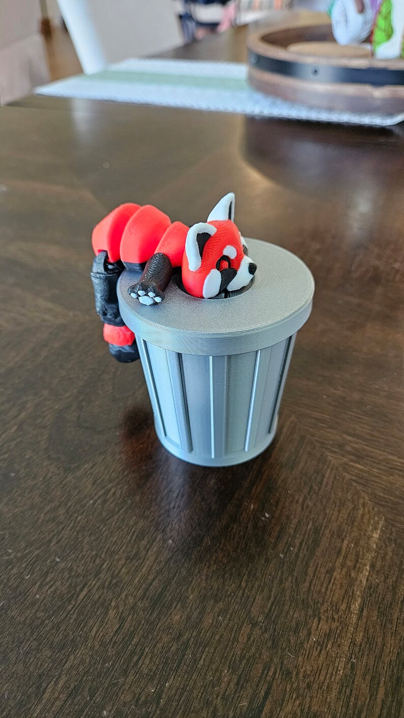 May include: A red panda figurine with white accents rests on a small, gray trash can. The panda has a black nose and paws. The trash can has vertical lines and a flat lid. The figurine is a whimsical decorative item.