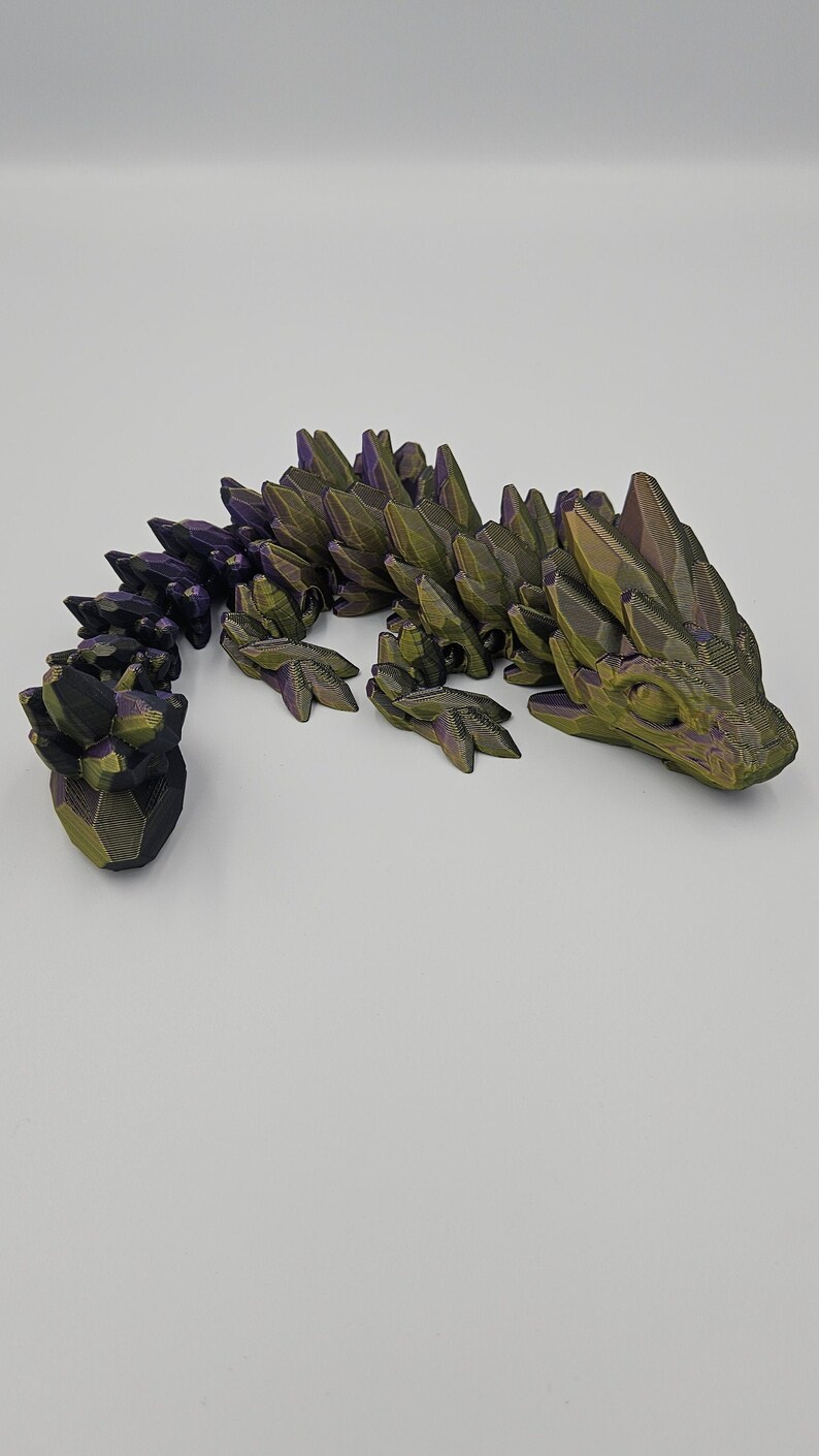 K&ouml;nnte beinhalten: Eine 3D-gedruckte Drachenfigur mit einem gr&uuml;n-violetten, irisierenden Finish. Der Drache ist in einer kriechenden Position mit erhobenem Kopf und eingerolltem Schwanz dargestellt.