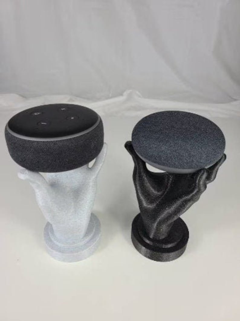Op de afbeelding: Twee handvormige standaards in zwart en grijs met een ronde bovenkant. Elke standaard houdt een zwarte en grijze slimme speaker.