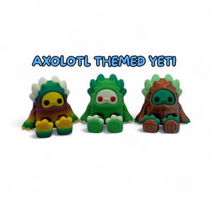 Puede incluir: Tres figuras de Yeti con tem&aacute;tica de ajolote. Las figuras son de colores arco&iacute;ris, verde y marr&oacute;n. Cada una tiene una cara redonda con dos ojos negros y una boca blanca o roja. El texto "AXOLOTL THEMED YETI" est&aacute; encima de las figuras.