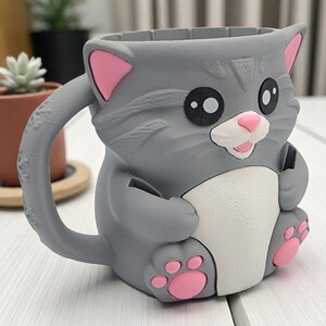 May include: A gray cat-shaped mug with pink accents on the ears and paws. The mug has a white belly and a smiling face. The handle is curved and attached to the side of the mug. The mug is made of a hard material.