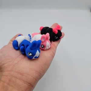 K&ouml;nnte beinhalten: Drei kleine Pl&uuml;schtiere in Form von Hasen. Der Hase links ist blau und wei&szlig;, der mittlere Hase ist rosa und wei&szlig;, und der Hase rechts ist schwarz mit rosa Ohren.