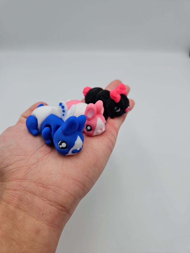 K&ouml;nnte beinhalten: Drei kleine Pl&uuml;schtiere in Form von Hasen. Der Hase links ist blau und wei&szlig;, der mittlere Hase ist rosa und wei&szlig;, und der Hase rechts ist schwarz mit rosa Ohren.