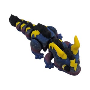 K&ouml;nnte beinhalten: Ein 3D-gedrucktes, bewegliches Drachenspielzeug im Galaxiethema. Der Drache hat einen blauen K&ouml;rper mit lila und wei&szlig;en Sprenkeln, schwarzen Akzenten und gelben Blitzmustern. Der Drache hat gelbe H&ouml;rner und einen schwarzen R&uuml;cken.